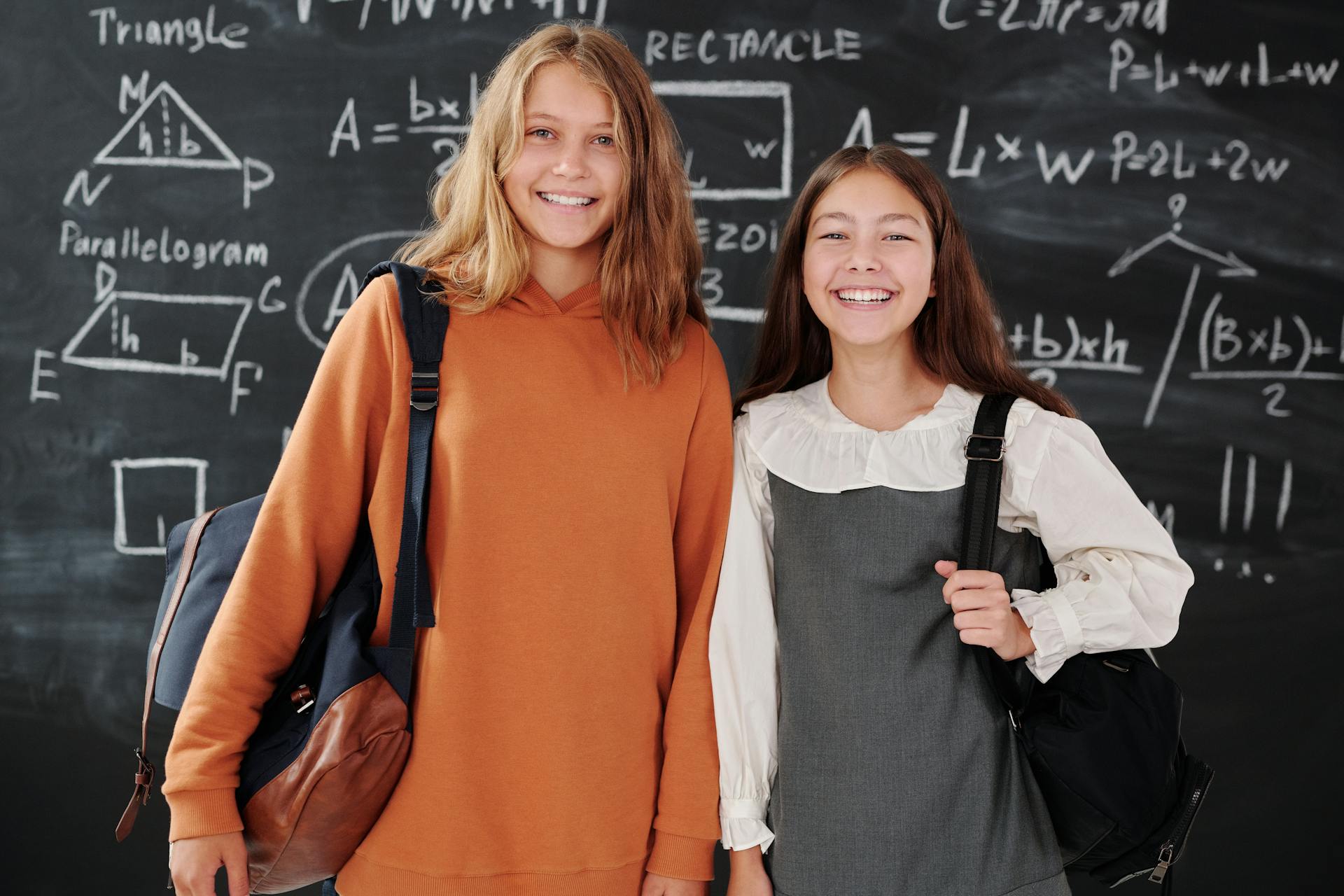 Two smiling students with backpacks standing in front of a chalkboard filled with math formulas.