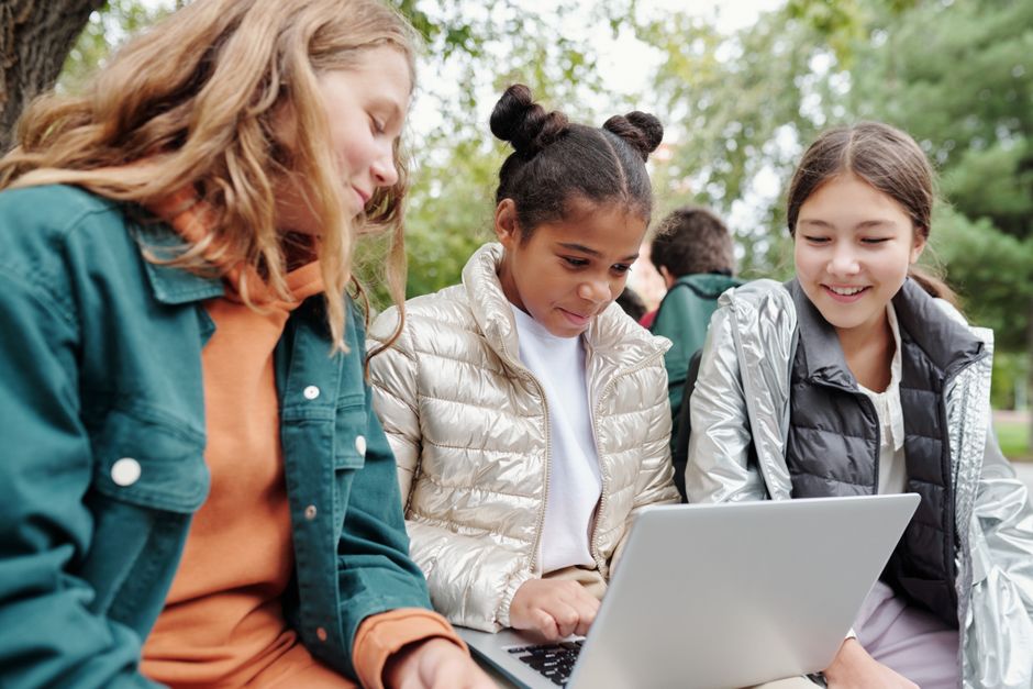 Three girls sitting together outdoors, smiling and learning on a laptop in a park setting.