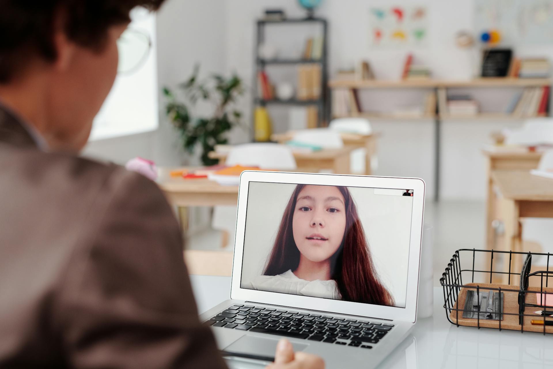 Teacher using laptop for virtual lesson with female student on screen in classroom setting.