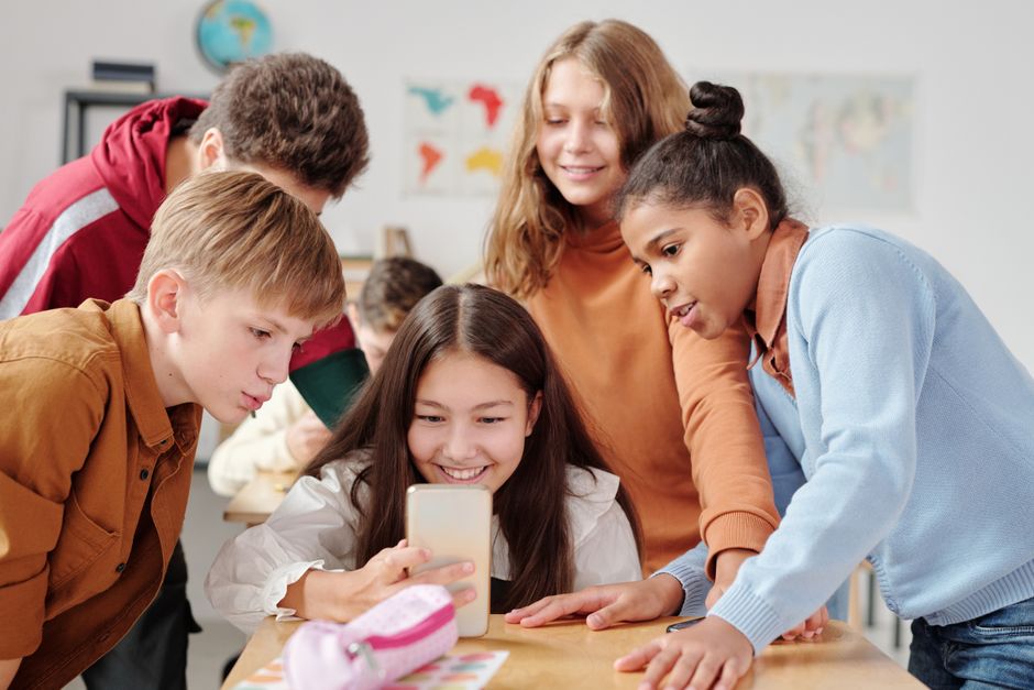 Group of teenagers in a classroom setting happily gathered around a smartphone.