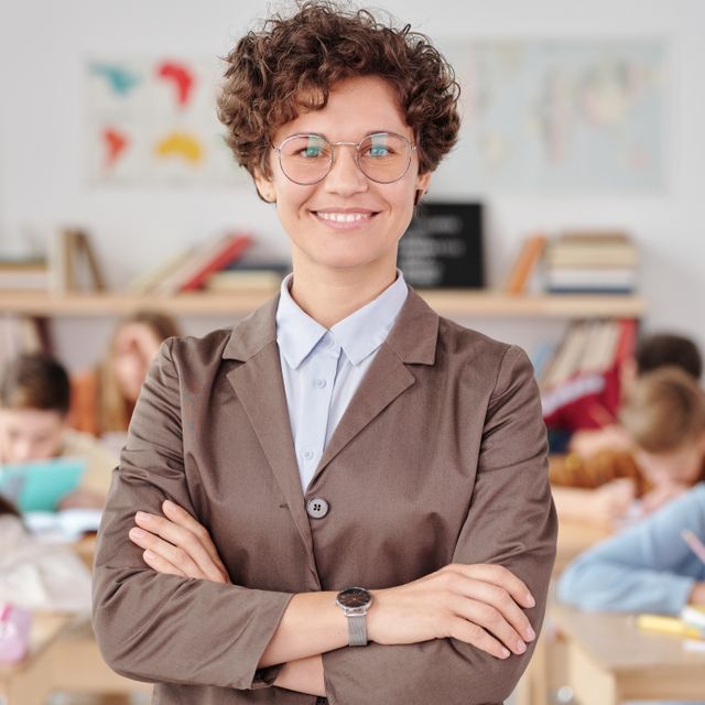 Lächelnde Lehrerin mit Brille steht mit verschränkten Armen in ihrem Klassenzimmer