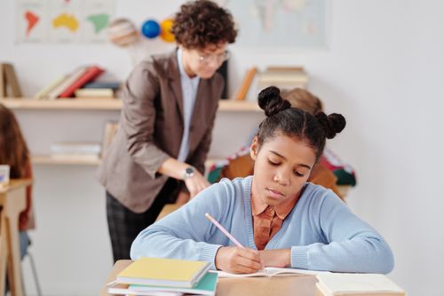 Free Focused young student writing during a classroom activity with teacher in background. Stock Photo