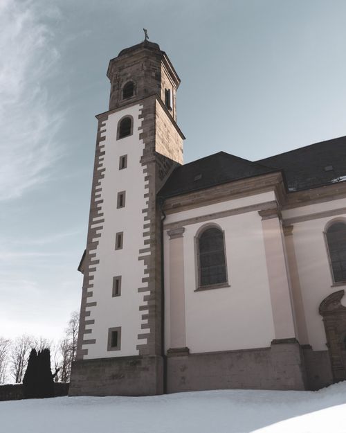 Free Gothic-style church tower in a snowy German landscape, surrounded by leafless trees under clear winter skies. Stock Photo