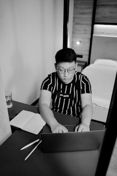 Asian man with eyeglasses typing on laptop in a modern minimalistic bedroom, black and white theme.