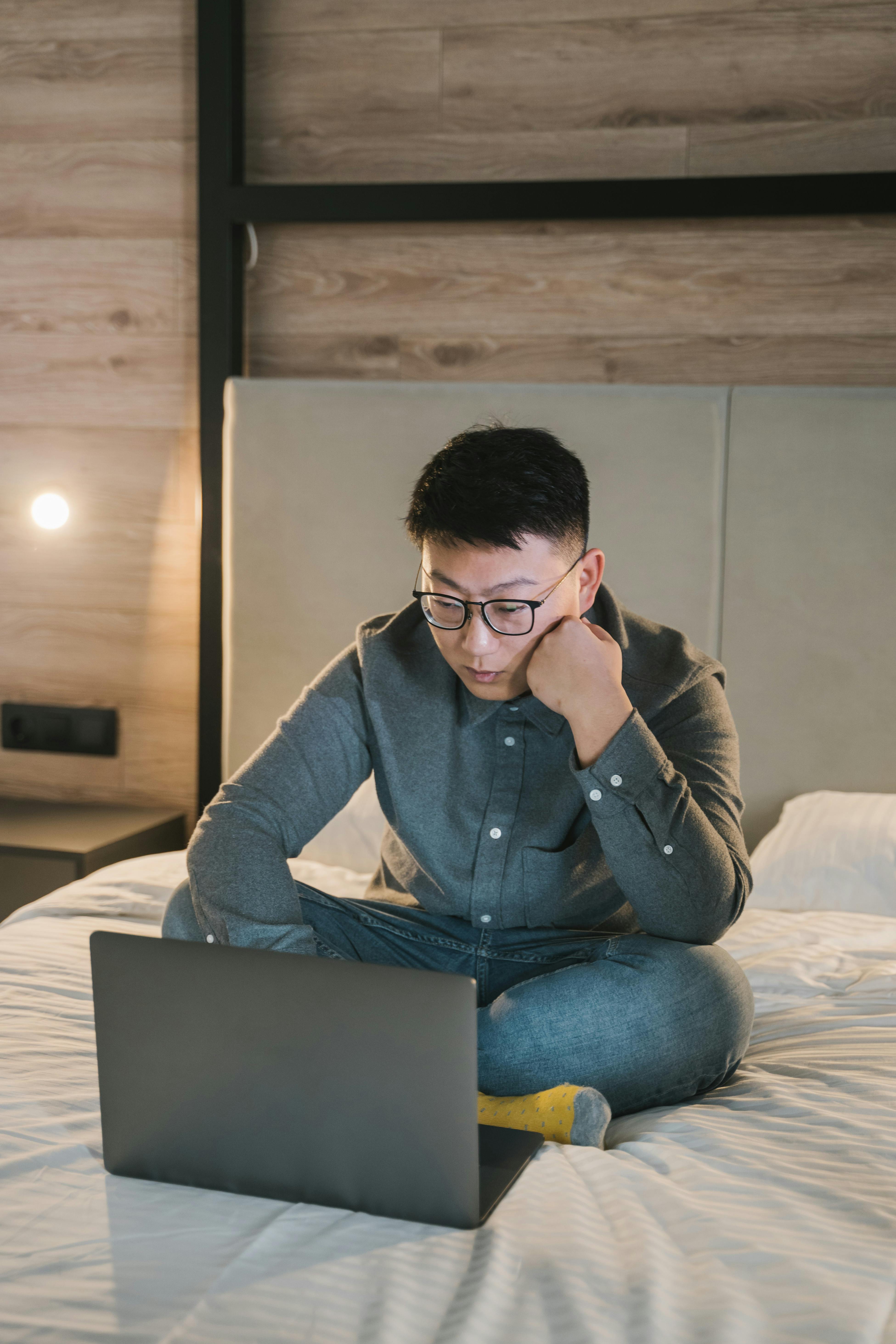Asian man working on a laptop in a cozy bedroom setting, focused on online tasks.