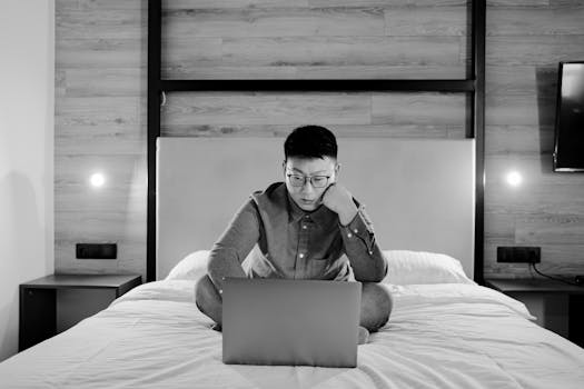 Asian man sitting on a bed working on a laptop in a grayscale home office setting.