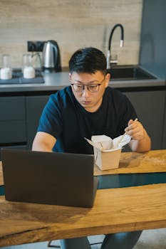 Asian man working on a laptop while eating lunch in a modern kitchen setting. Remote work lifestyle.