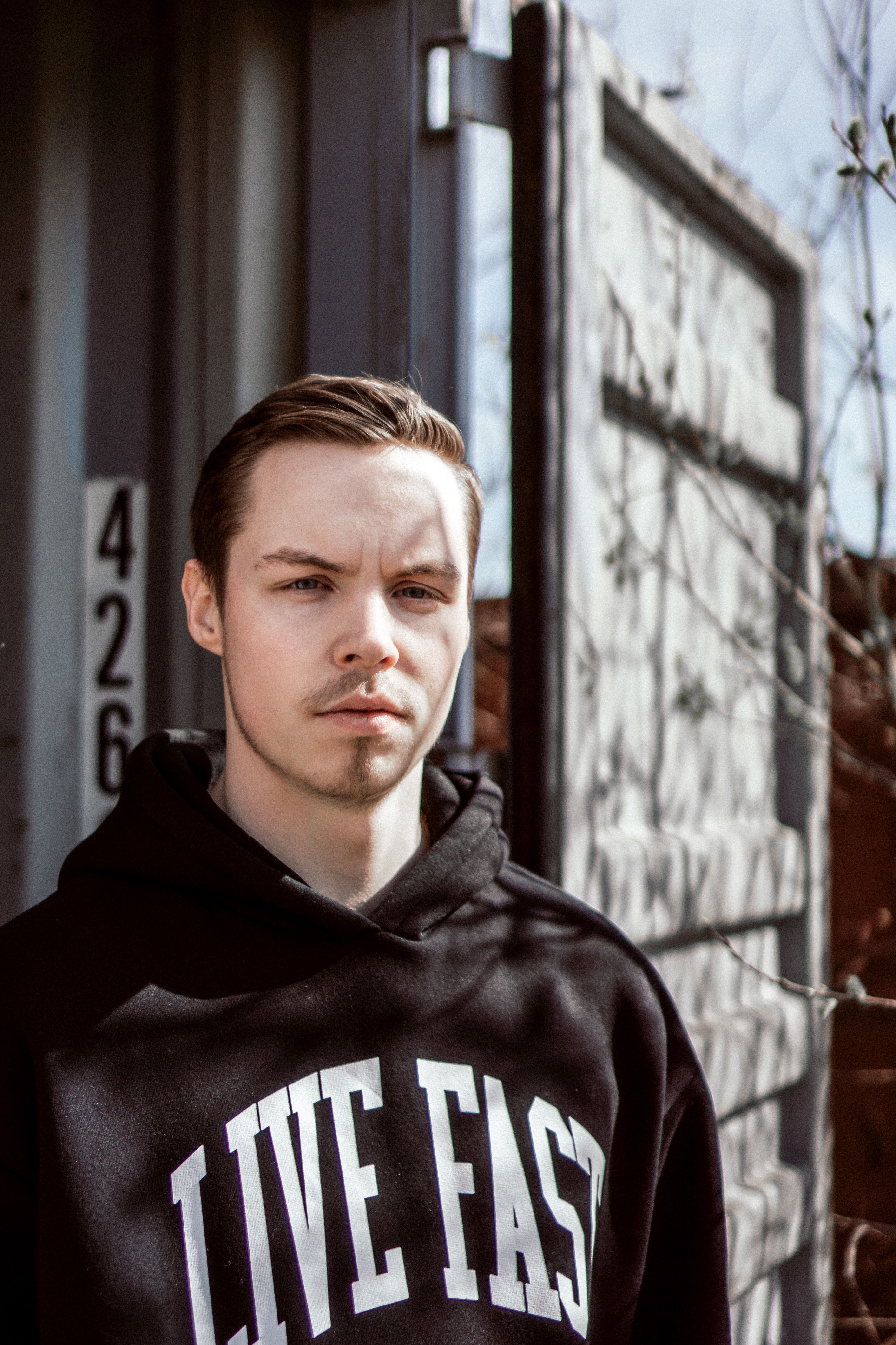 Portrait of a young man in a black hoodie with 'Live Fast' text, standing near a cargo container outdoors.