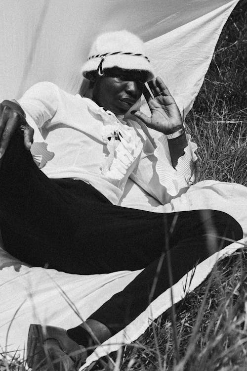 Black and white of African American serious man in elegant blouse and panama hat resting on sheet in meadow