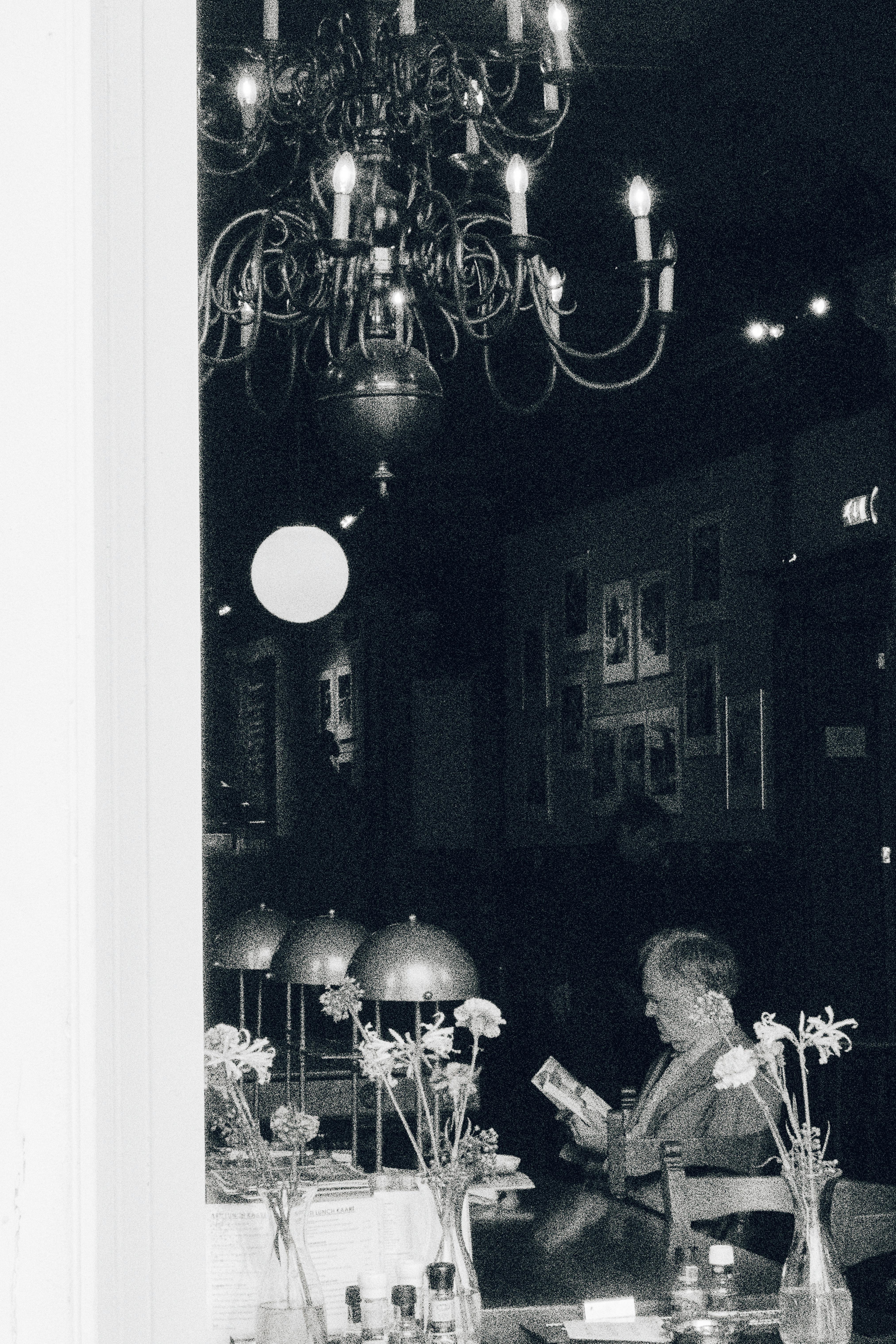 Black and white image of a person reading in a cozy café beneath a grand chandelier.