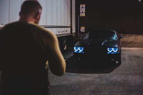 Back view of unrecognizable male on parking lot standing near modern black automobile with blue headlights parked at night