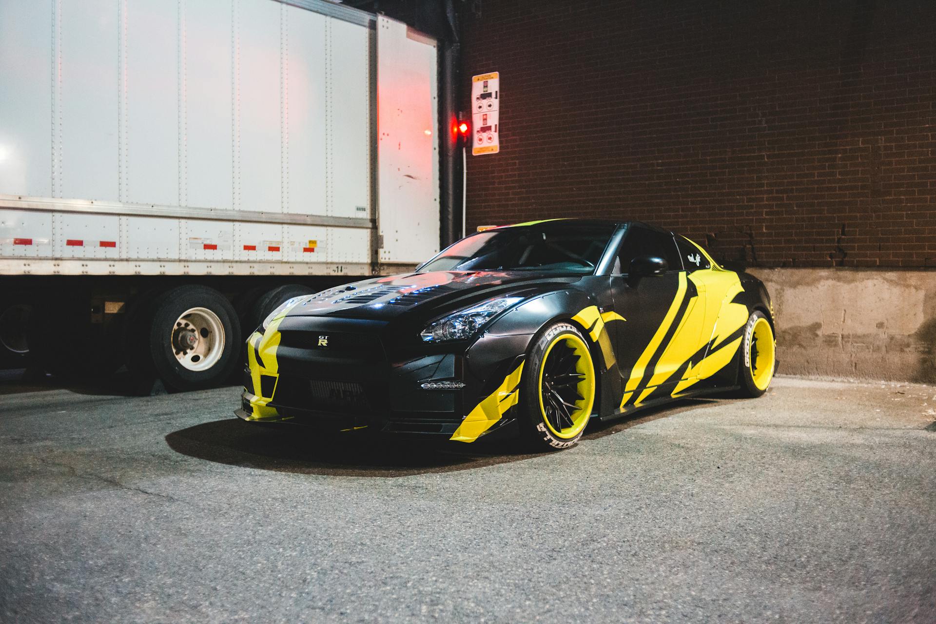 Custom sports car in vibrant yellow and black wrap parked by a brick wall at night.