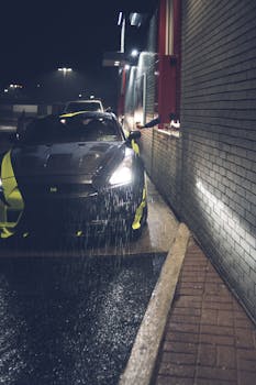 一辆带有黄色条纹和前灯的当代黑色汽车，在雨夜停靠在柏油路上，旁边是一栋有砖墙的建筑