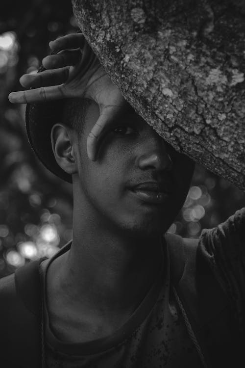 Black and white thoughtful young African American male standing behind tree and thinking while looking at camera