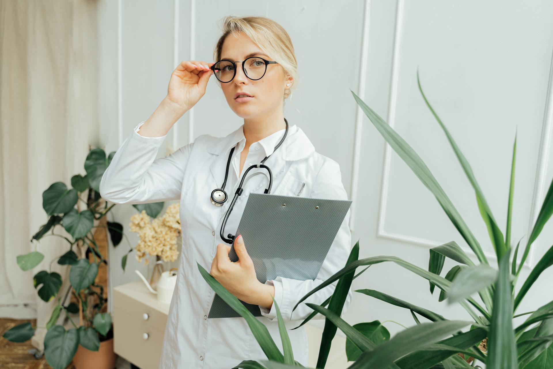 Confident female doctor wearing eyeglasses and holding a clipboard in an indoor office setting.