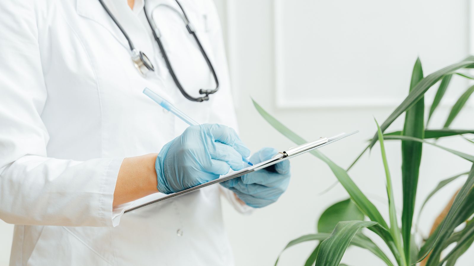 A healthcare professional wearing gloves takes notes on a clipboard with a stethoscope around their neck.