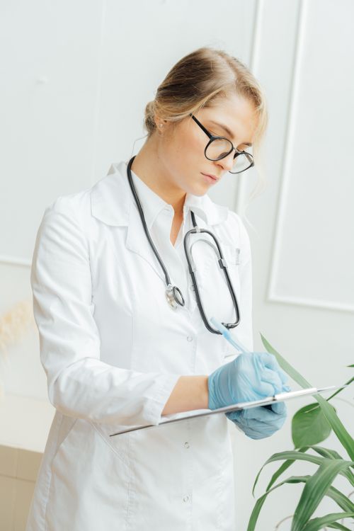 Free Female doctor with eyeglasses taking notes in a hospital, wearing a stethoscope and gloves. Stock Photo