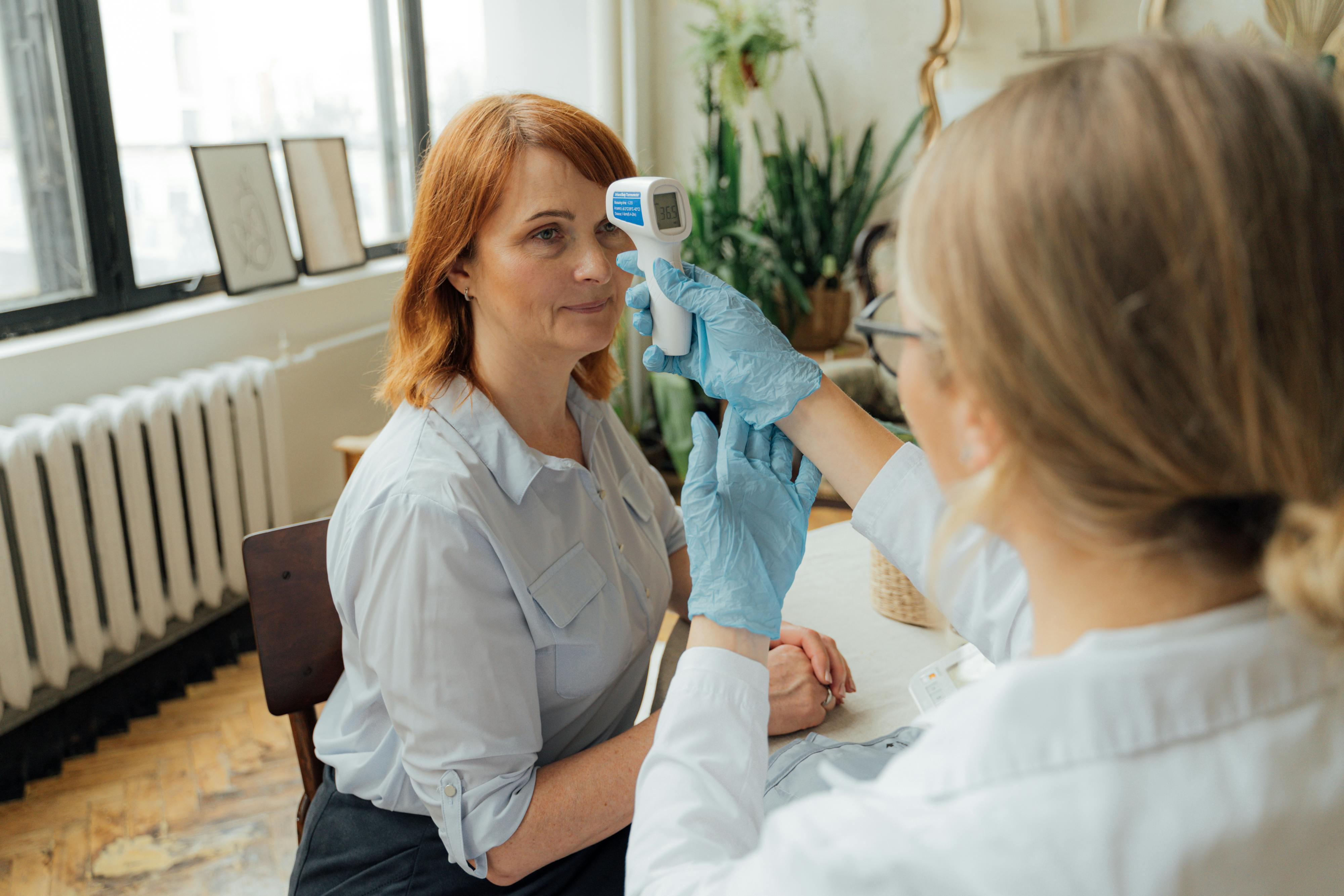 A healthcare worker measures a patient's temperature indoors using a digital thermometer.