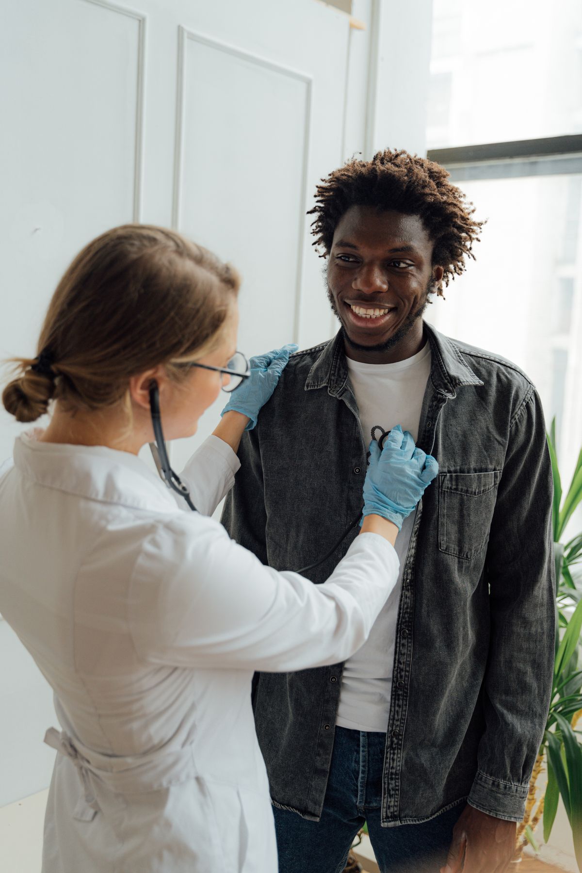 A clinician in a white coat and gloves listening to a smiling man's chest with a stethoscope.
