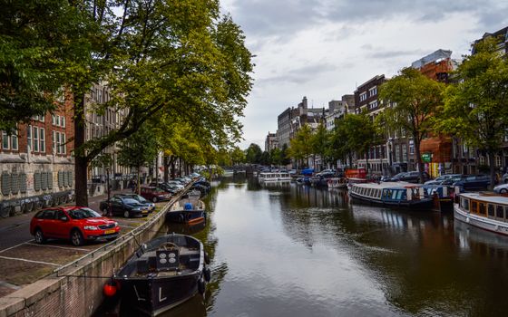 Various cruise boats moored on canal flowing in aged city between typical buildings against cloudy sky