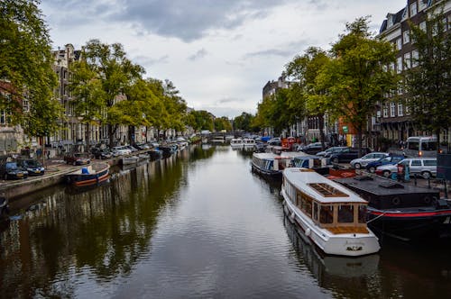 Picturesque scenery of modern and aged cruise boats moored on canal flowing between typical buildings against cloudy sky in Amsterdam