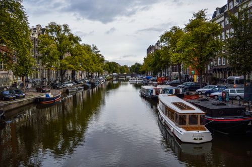 Free Picturesque scenery of modern and aged cruise boats moored on canal flowing between typical buildings against cloudy sky in Amsterdam Stock Photo