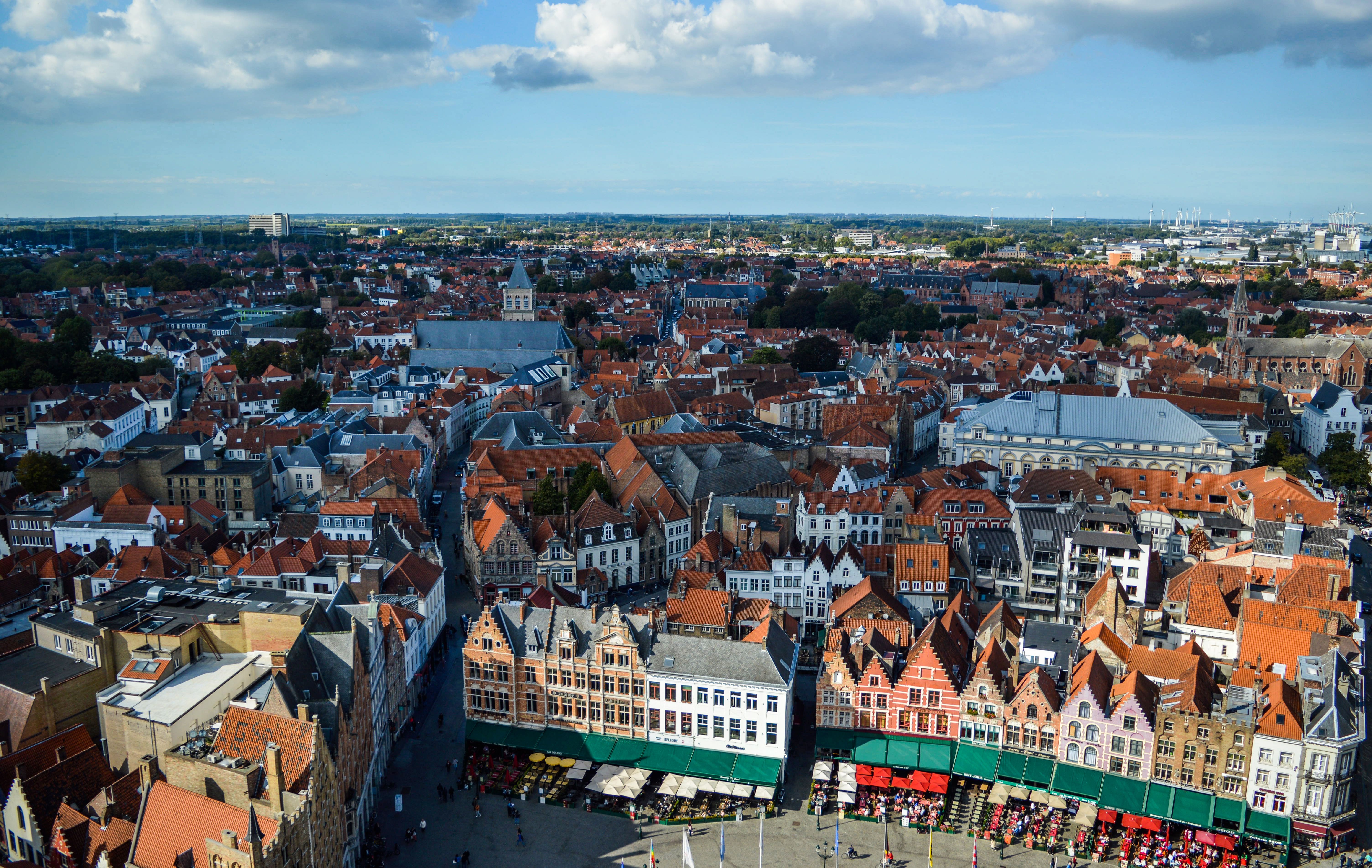Scenic drone view of Bruges Market Square surrounded by aged colorful buildings against cloudy blue sky