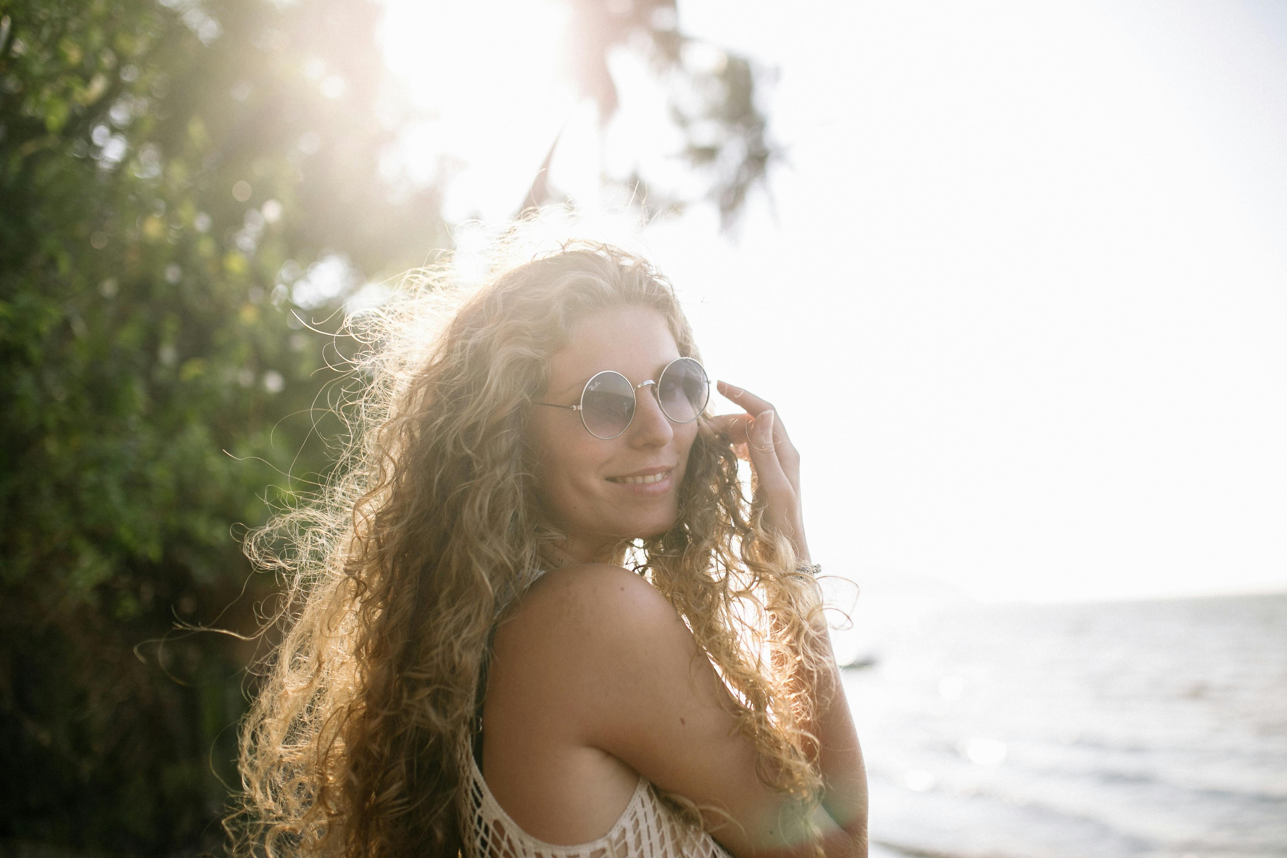 一位留着卷发、戴着墨镜的年轻女子在海边享受阳光明媚的一天，散发着幸福和无忧无虑的气息