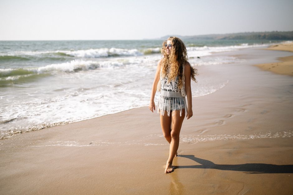 A woman with curly hair walks barefoot along a sunny beach, enjoying the waves.