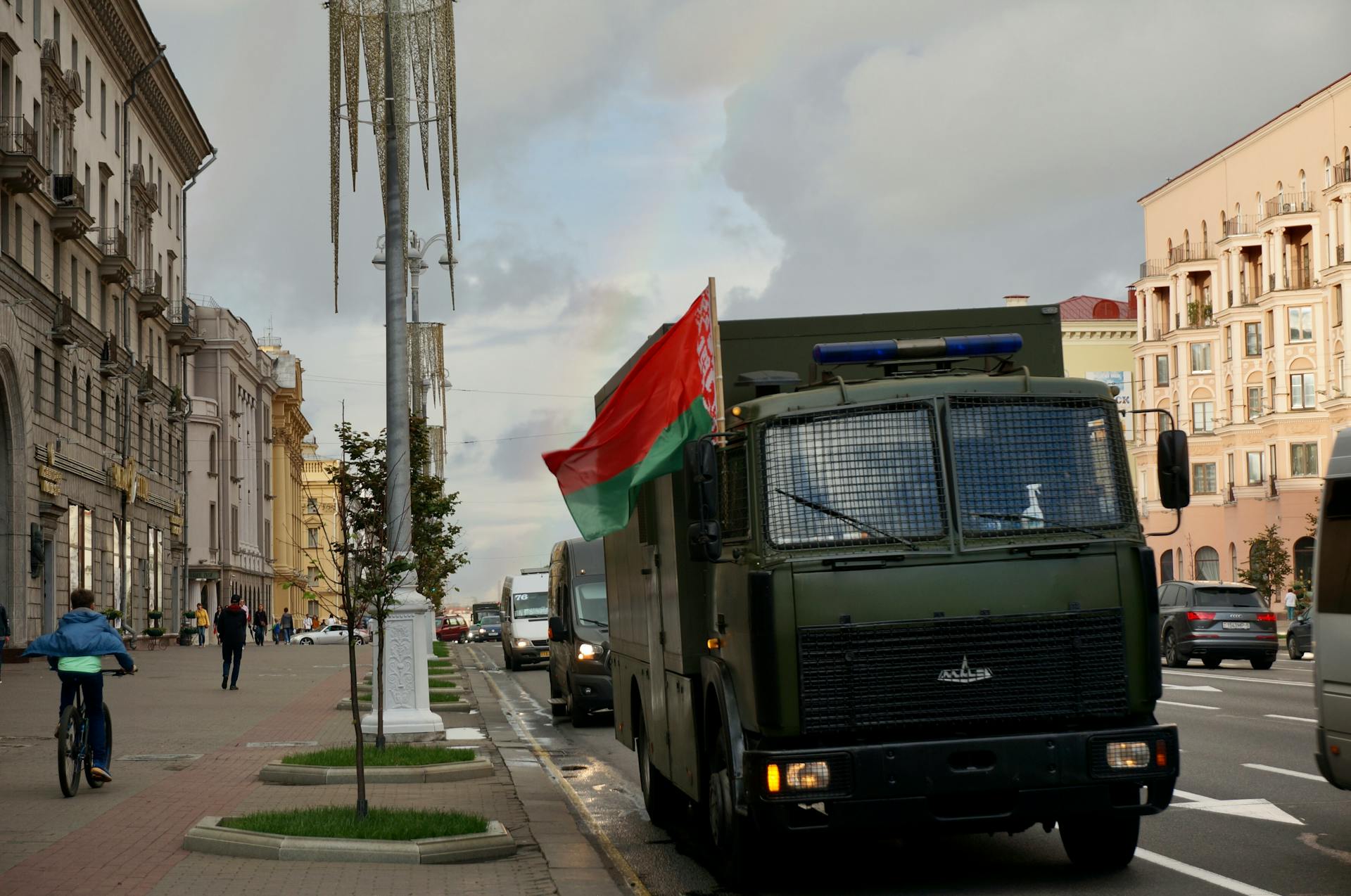An armored truck carrying a flag drives down a bustling urban street with historic architecture.