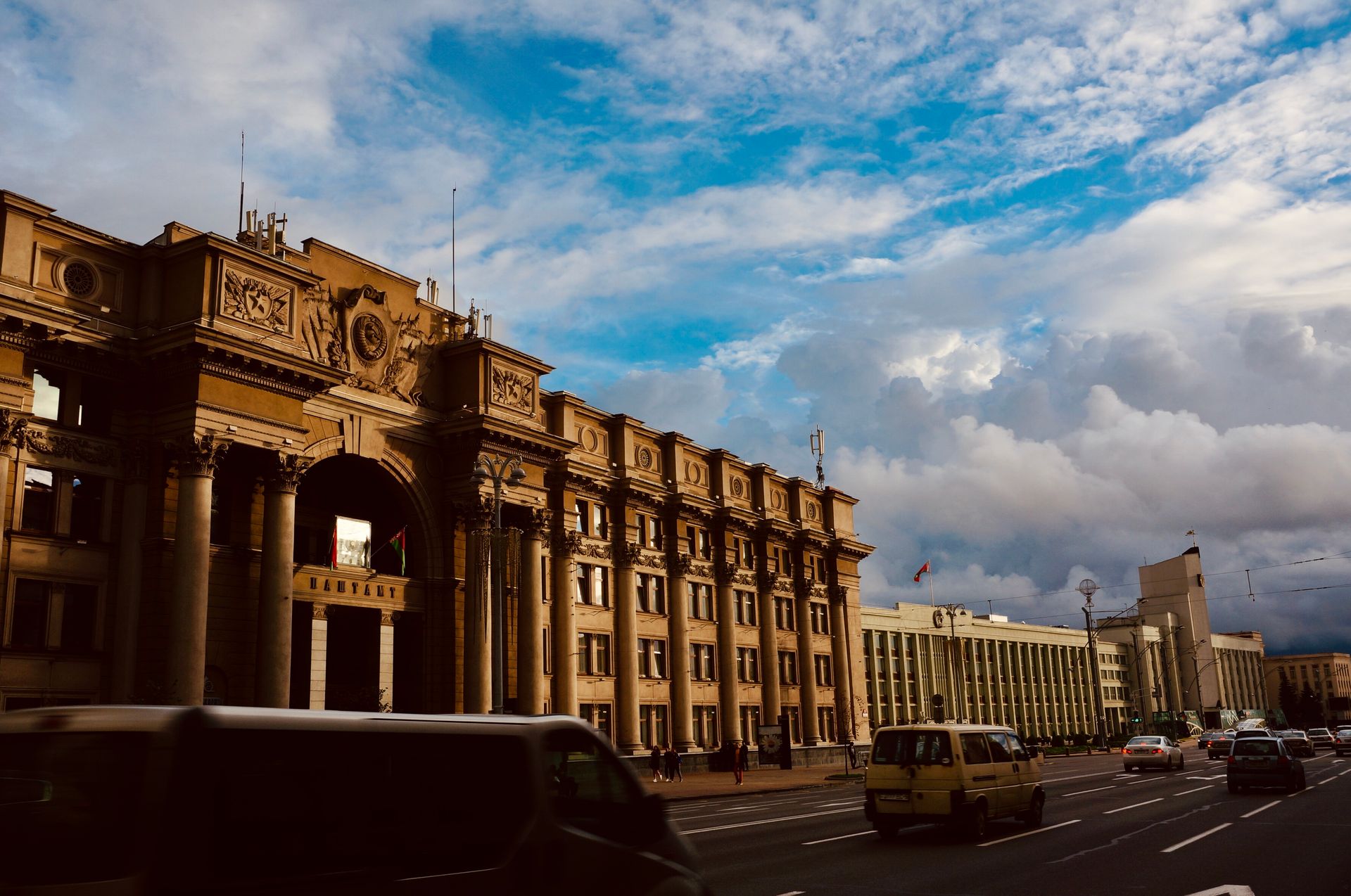 A grand government building in Minsk, Belarus, set against a dynamic cloudy sky with road traffic below.