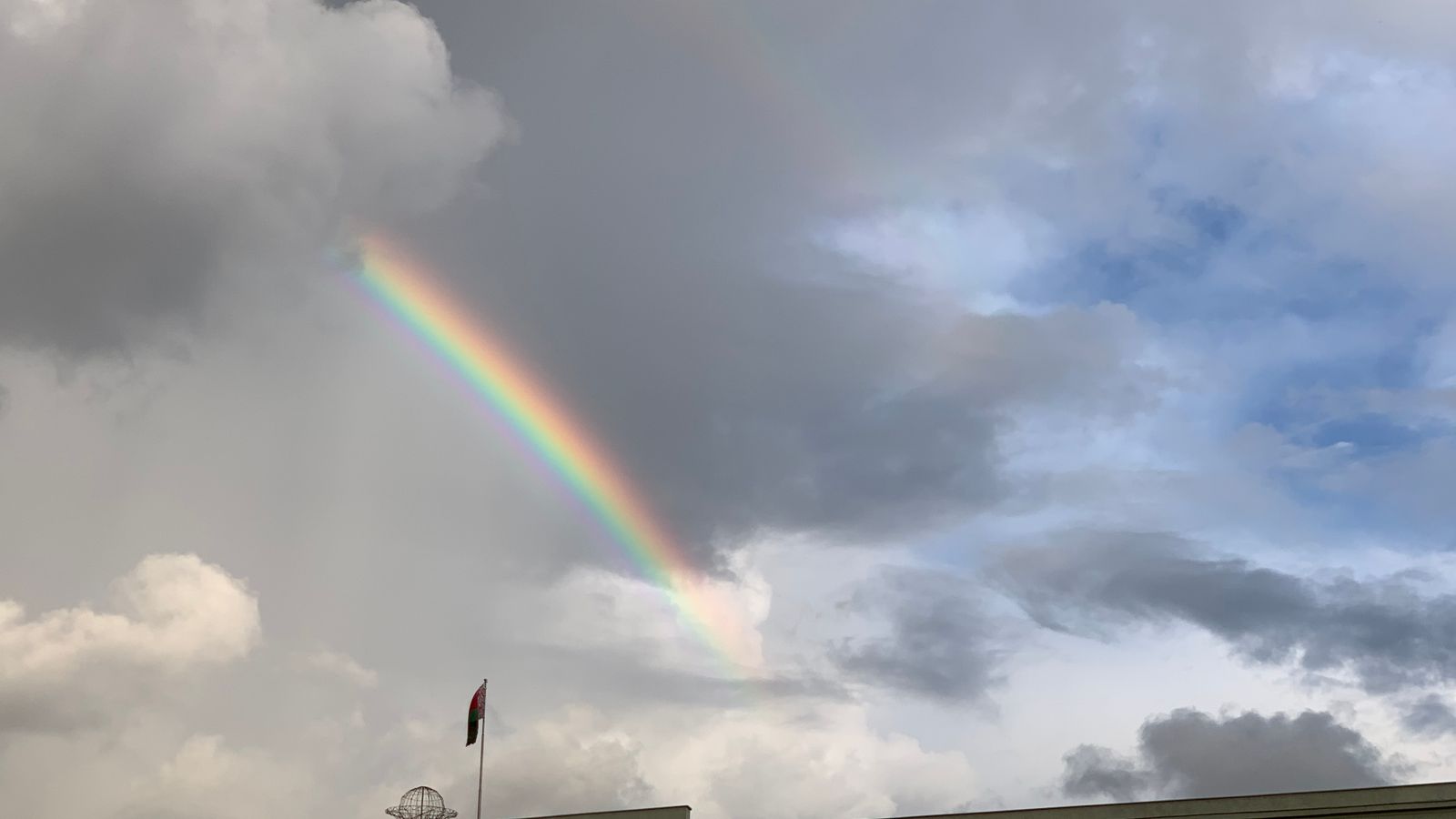 Vibrant rainbow arches over a building with a national flag under a cloudy sky.