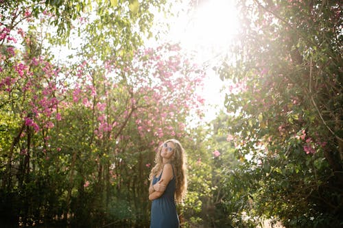 Woman in Blue Dress Wearing Sunglasses Posing near Trees with Pink Flowers