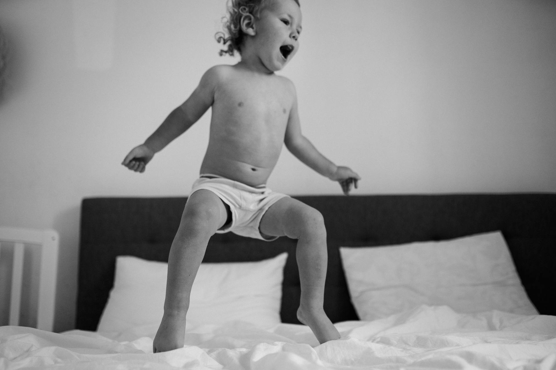 Grayscale image of a shirtless child energetically jumping on a bed indoors.