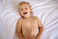 一个快乐的孩子躺在室内干净的白色床上笑着，营造出温暖俏皮的氛围