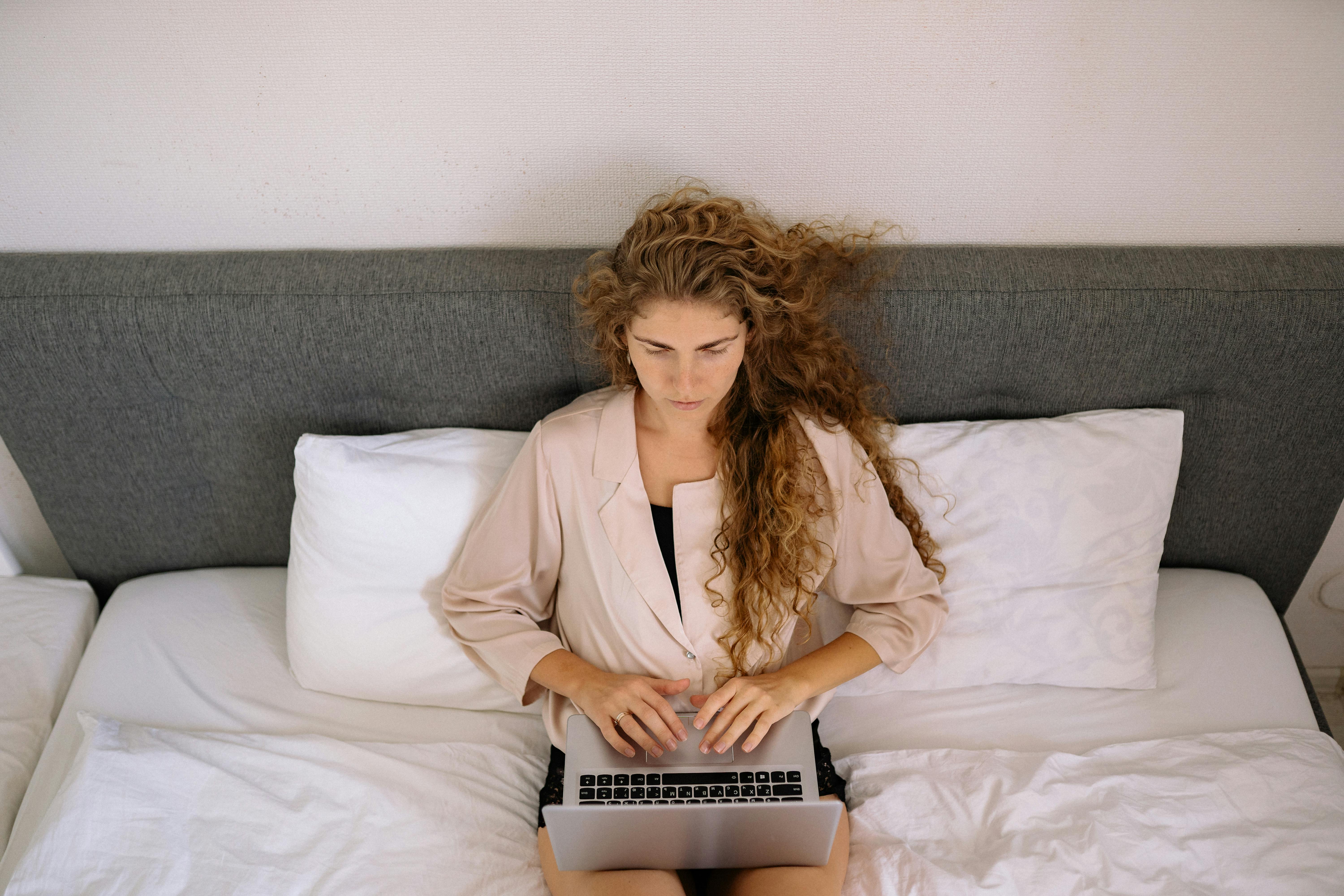 A woman with curly hair types on a laptop while sitting on a bed.