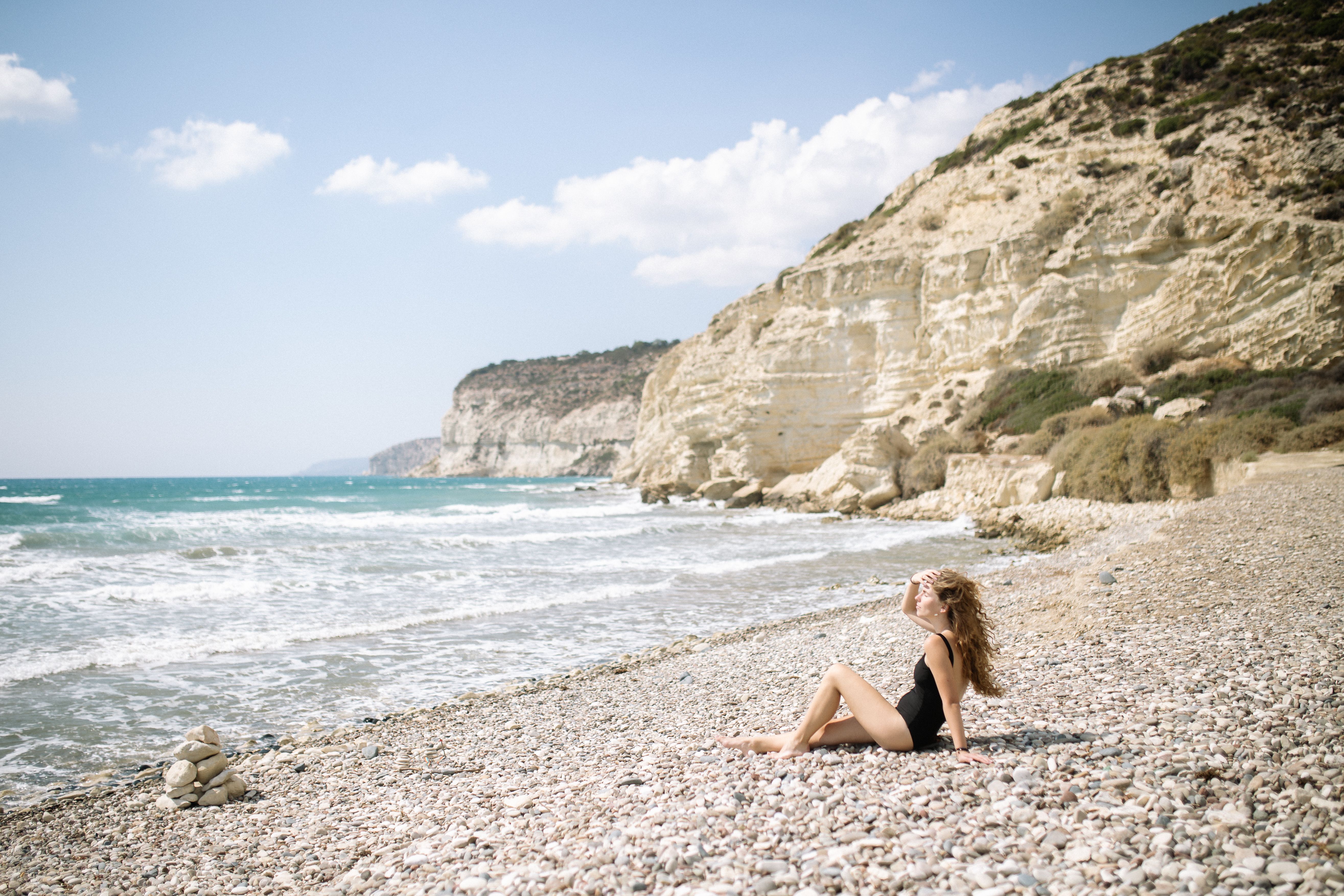 A woman in a black swimsuit relaxes on a pebble beach by an ocean under clear skies. - Photo by Yan Krukau on Pexels