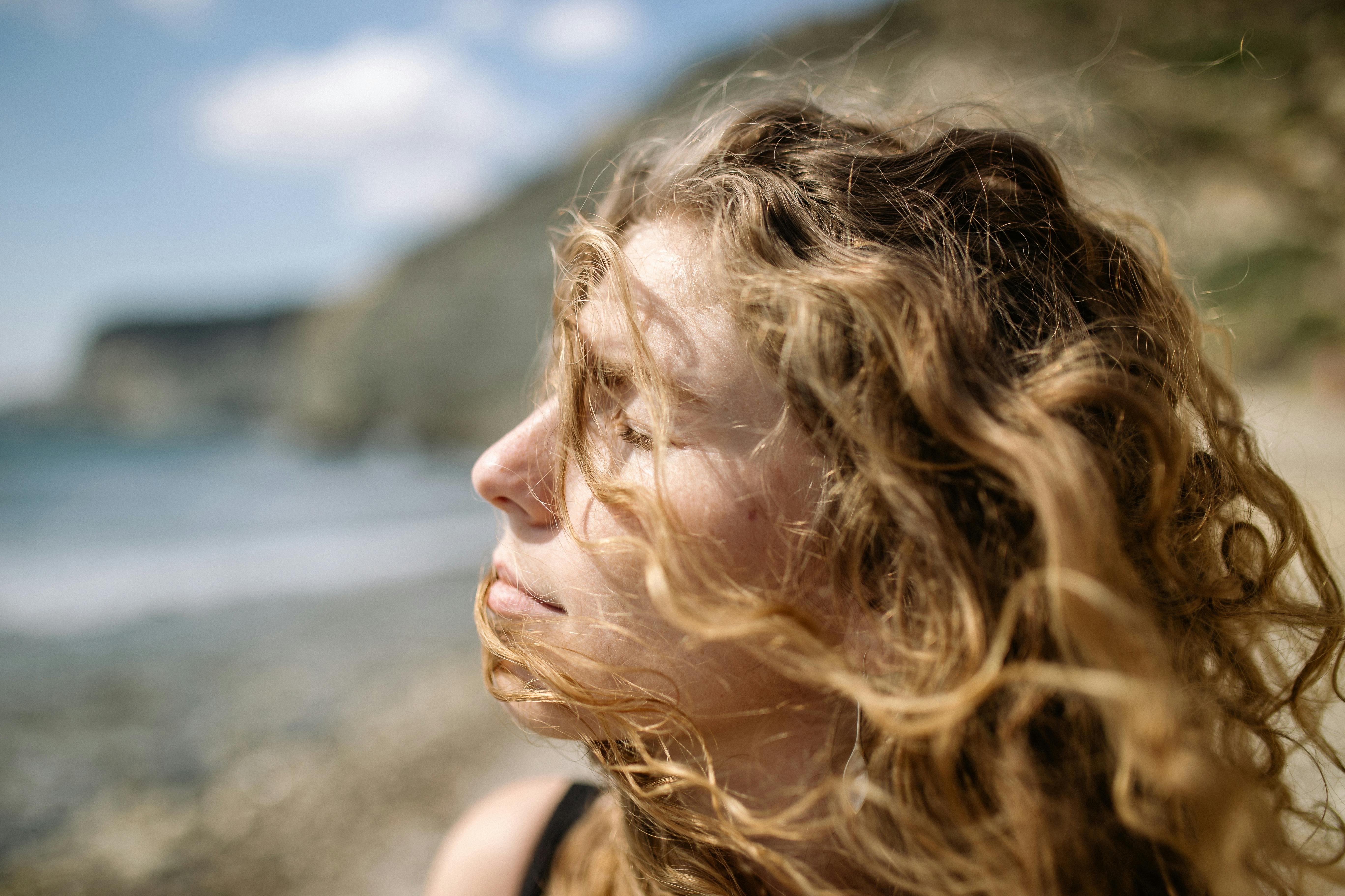 Profile of a woman with curly blonde hair, eyes closed, enjoying a windy day at the beach.