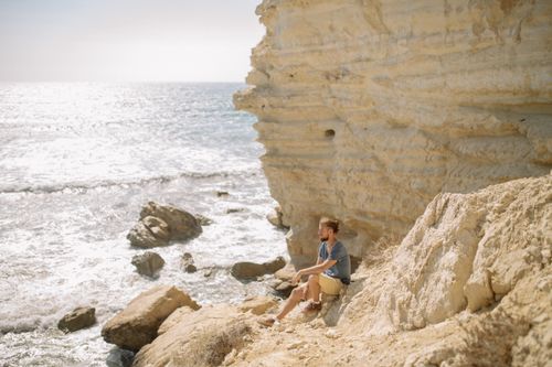 Free A man enjoys a peaceful moment sitting by a rocky seaside cliff under the bright sun. Stock Photo