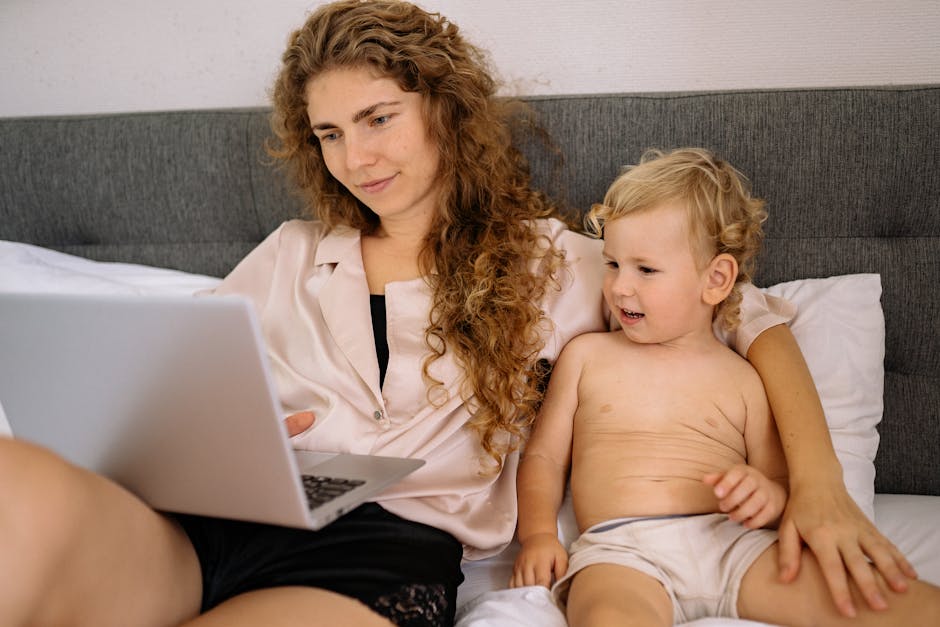 A mother with curly hair and her toddler son enjoying time together on a bed while using a laptop.