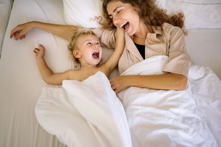How Toddler Beds Ease Sleep Transitions for Busy Parents A joyful mother and child laughing together in a cozy indoor setting, wrapped in white blankets.