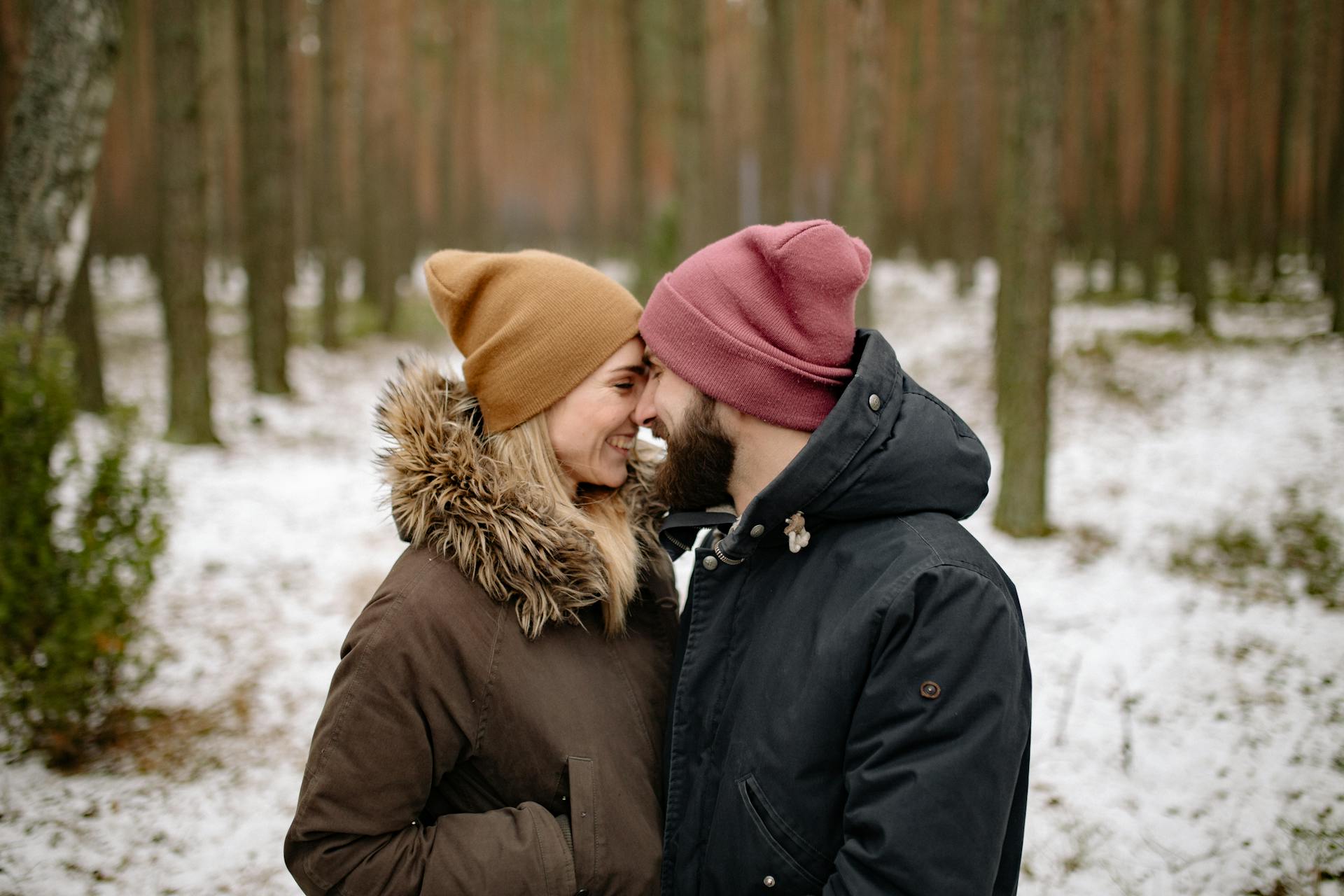 A happy couple in warm jackets and beanies share a loving moment face to face in a winter forest.