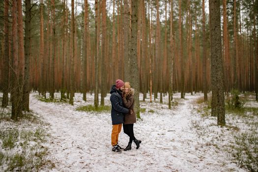 A couple wearing winter coats and beanies embraces in a snow-covered forest.
