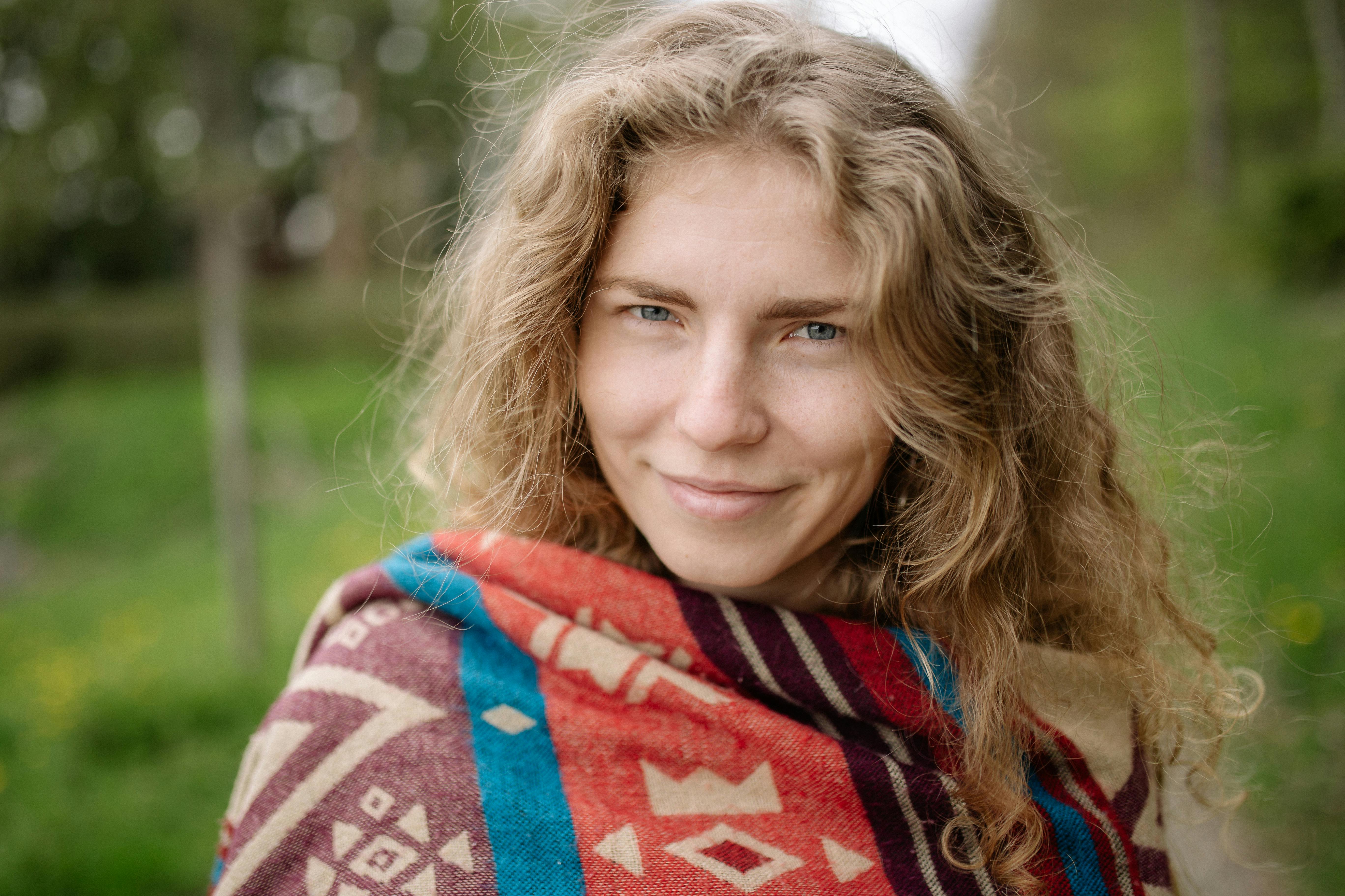 一位开朗的金发女子，裹着彩色毯子，在自然环境中展露温暖笑容