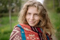 一位开朗的金发女子，裹着彩色毯子，在自然环境中展露温暖笑容