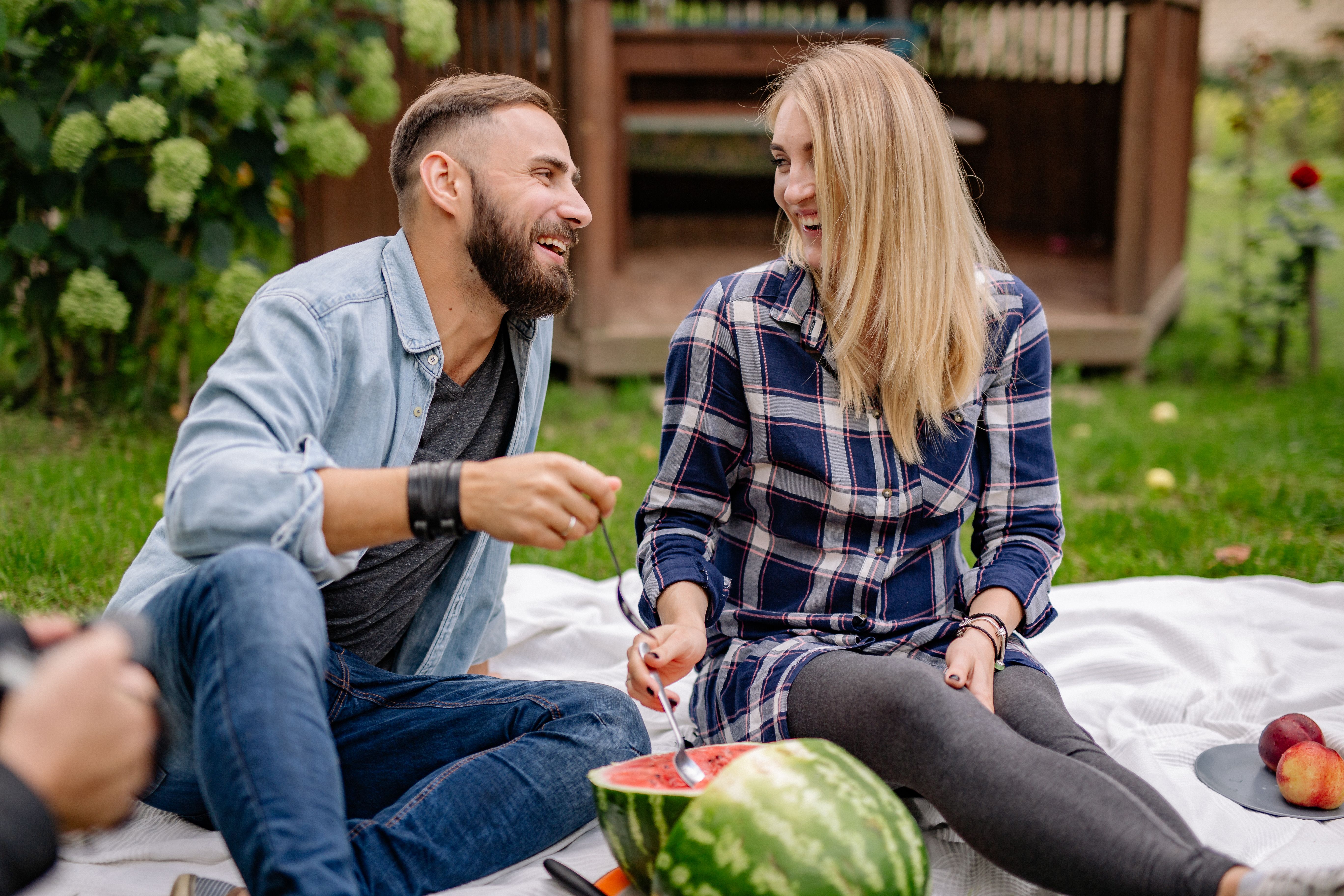 Casal sorridente curtindo um piquenique ao ar livre em um jardim, com melancia e frutas ao redor.