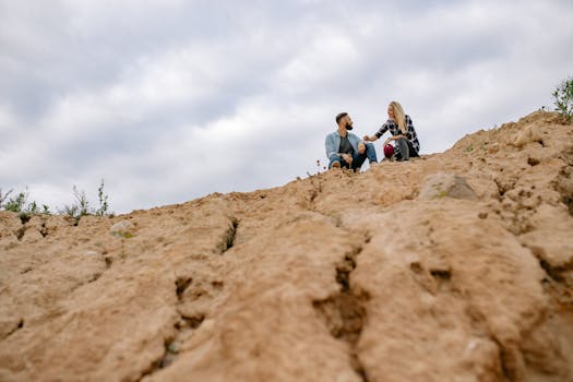 A couple enjoys a calm moment sitting on a rocky cliff in a desert setting, under a cloudy sky.