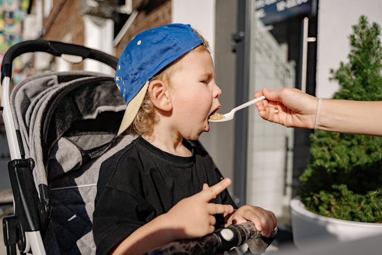 Mother Feeding Her Baby Son Sitting In A Stroller Outdoors 