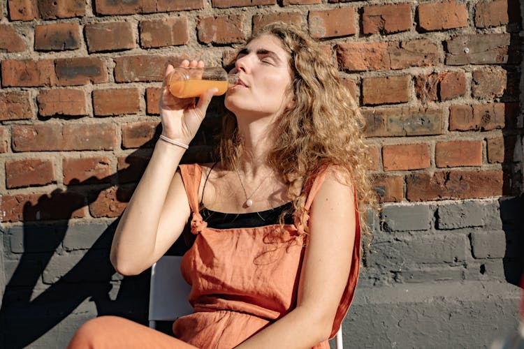 Woman Leaning On A Brick Wall And Drinking Juice Out Of A Bottle