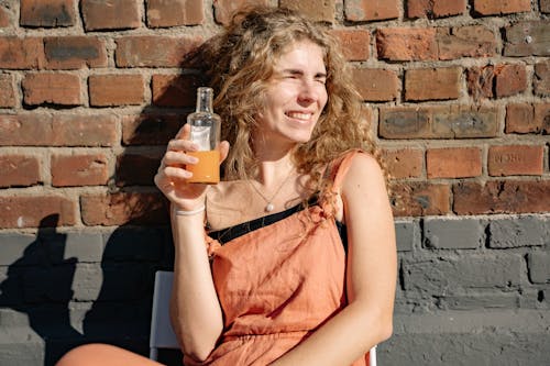 Woman in Orange Jumper Sitting Down while Holding Clear Glass Bottle with Orange Liquid